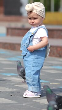 Curious toddler girl in a fashionable denim outfit with decorative wings exploring a city square, surrounded by a flock of pigeons walking on the pavement on a bright sunny summer day