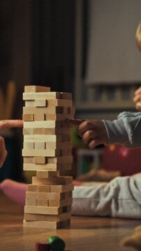 A mother and her young daughter sit on a carpeted living-room floor, arranging bright wooden Jenga blocks into a wobbly tower, forming a family play scene focused on learning and entertainment