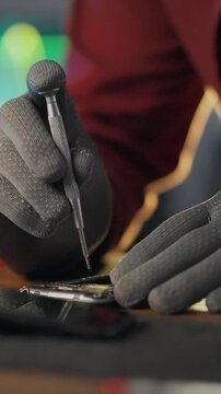 An electronics mechanic in a workshop repair shop grips a screwdriver, unscrewing components while diagnostic equipment runs, performing precise technical work in a focused, industrial environment