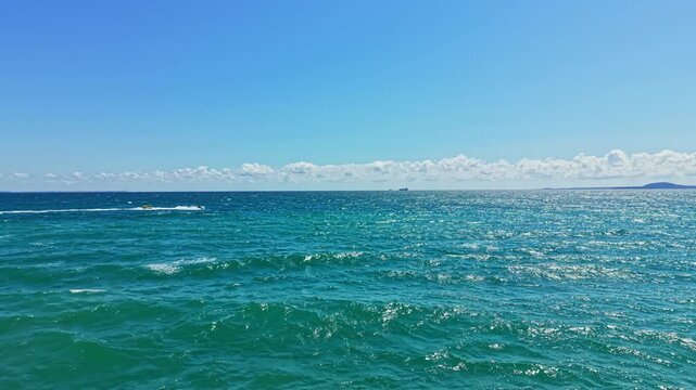 Waves gently roll on the surface of the ocean under a bright blue sky. Clouds linger in the distance while a ship can be seen far off at sea.