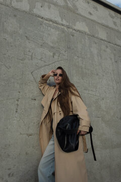 Stylish young woman with long hair wearing a beige trench coat, sunglasses, and holding a black leather bag, posing against a textured grey concrete wall in an urban street style fashion shoot.