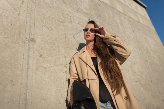 Trendy shot of a confident young woman with long hair wearing a beige trench coat and adjusting her sunglasses against a concrete wall under a blue sky. Urban fashion street style.