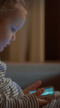 Child sits on sofa in the evening, eyes fixed on a smartphone displaying bright cartoons, the scene set in a living room, evening ambience, everyday home media activity. Soft lamp light casts shadows