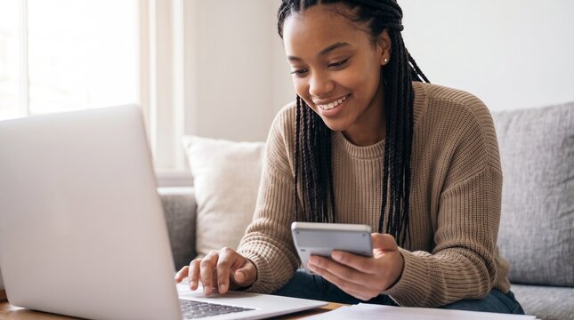 Smiling young African American woman calculating home budget and applying for mortgage loan online using laptop and calculator while sitting on sofa in modern cozy living room indoors.
