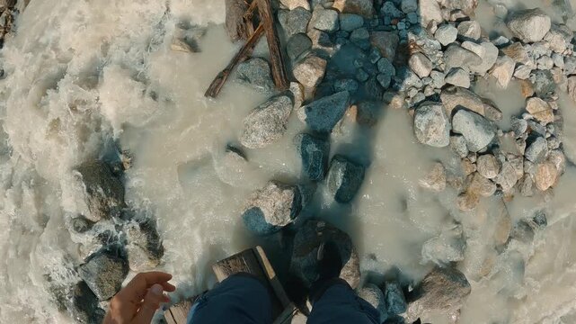 First person view of hiker stepping across rustic wooden bridge spanning turbulent mountain stream. Sunlit scene with white water foam, gray rocks, blue pants, hiking boots