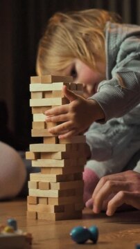 Parents and their young daughter sit on the living-room floor at home, arranging block tower board game together, the scene set in a quiet evening atmosphere with soft lighting casting gentle shadows
