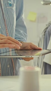 Seamstress in tailor's workshop uses a tablet while surrounded by hanging garments and fabric rolls, the bright lighting highlighting the space as she updates inventory and prepares orders for retail