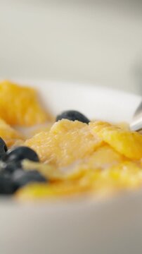 Steel spoon in kitchen scoops yellow cornflakes and fresh blueberries onto bowl, depicting breakfast preparation step that emphasizes simple, healthy morning meal, served in a bright kitchen setting