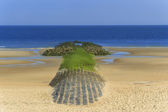 a breakwater stone path crosses the Ostend sand toward the calm sea, its moss‑touched surface meeting the gentle water under a partly bright sky.