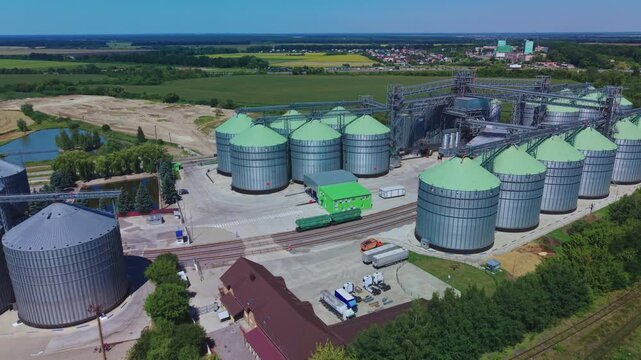 Dynamic aerial overview reveals rows of silver and green metal silos near railway tracks and cargo cars at modern grain storage facility.