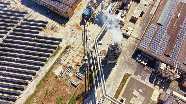 Aerial view shows an industrial facility with solar panels, pipes, and smoke. Equipment and structures are visible in an energy production zone during bright daylight.