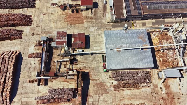Workers are operating machinery at a wood processing plant. Stacks of logs are placed in rows. The scene shows a busy industrial area where wood is prepared for use.