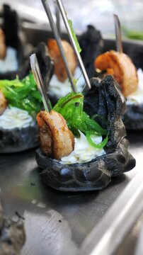 Chef decorating gourmet black tartlets with grilled shrimp, cream cheese, and chuka seaweed using tweezers