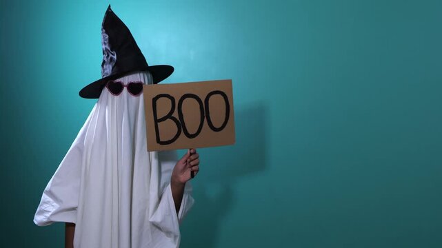 Ghost costume holding a BOO sign against a teal wall for Halloween