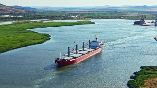 A red cargo ship moves along a river. Green fields line the banks. In the background, hills are visible under a clear sky. The ship is part of a trade route transporting goods.