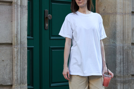 Woman in white t-shirt and beige shorts holding a pink drink outside a green door