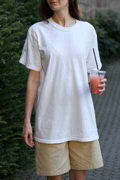 Woman in white t-shirt and beige shorts holding a pink drink outdoors
