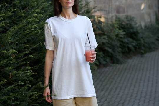 Woman in white t-shirt holding a pink beverage outdoors near green foliage