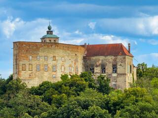 Hilltop castle amid trees. A castle stands on a hill, partially hidden by trees on a sunny day with blue skies and fluffy clouds. © Анна Терелюк