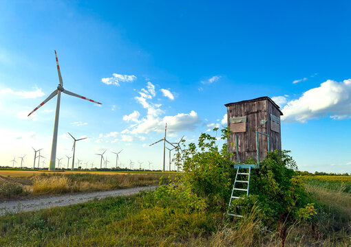 Turbines by wooden field structure. Wind turbines stand tall in a field next to a wooden structure during sunny weather. A dirt path leads to the scene.