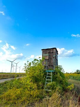 Old wooden structure near wind turbines. A wooden structure stands alone on a field. Wind turbines spin in the background under a blue sky.