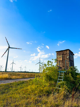 Wind turbines near a wooden structure. A wooden structure stands beside wind turbines in a field during daytime. The sun shines brightly in the blue sky.