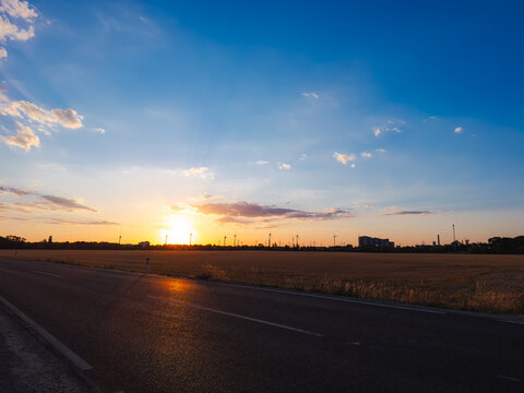 Sunset over a field by road. The sun sets over an open field next to a road. Clouds scatter in the sky as light fades.