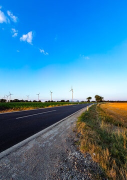 Evening light on windmills. A road runs alongside fields with windmills in the distance under a clear sky. The sun is setting in the evening.