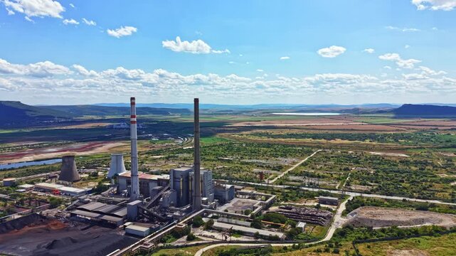 A large power plant stands in an industrial zone next to a river. Fields and hills surround the site. The sky has some clouds and looks clear. It is daytime.