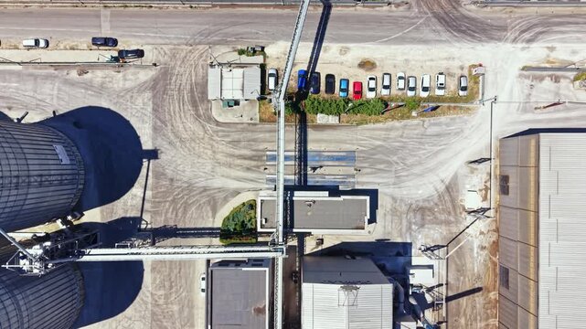 A look at an industrial area shows two large silos, several buildings, and a row of parked cars. The scene takes place under clear skies during daylight hours.