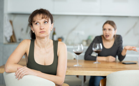 Sad middle-aged woman sitting at table and other woman brawling to her behind in the kitchen