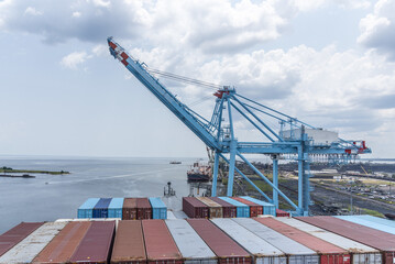 Naklejka premium Large container vessel alongside a busy container port with loading crane and stacked shipping containers, with visible entrance to the port of Mobile, Alabama, USA.
