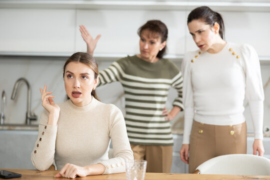 Upset middle-aged woman sitting at the kitchen table with her back to women quarreling angrily to her