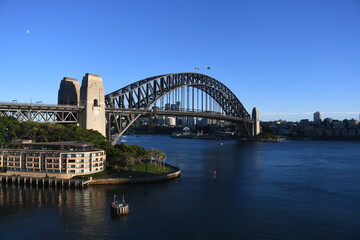 Naklejka premium Sydney Harbour Bridge spans the blue harbour waters under a clear sky, with residential buildings and parkland visible in the foreground.