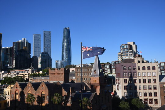 An Australian flag flies on a flagpole above historic brick buildings with modern skyscrapers visible against a clear blue sky in the background.