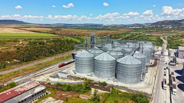 Several large metal silos are seen beside railway tracks in an agricultural area. Fields and hills are visible in the background under a clear sky.