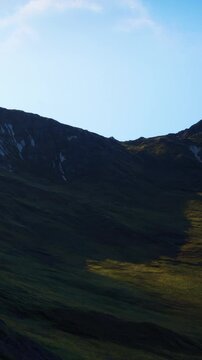 A mountain landscape is visible with steep slopes. The sun shines on green grass and creates shadows on the terrain. The scene shows a clear blue sky above the mountains.
