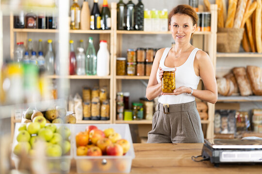 Adult female seller at counter offering olives in jar in grocery store