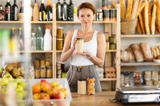 Adult female seller at counter offering beans in jar in grocery store