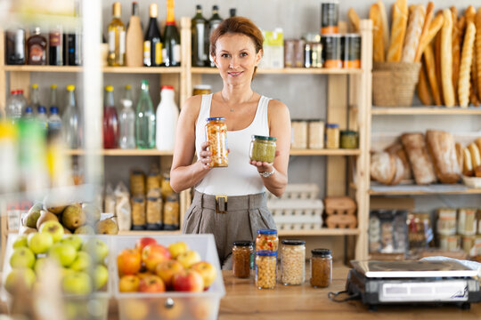 Adult female seller at counter offering green peas and beans in jar in grocery store