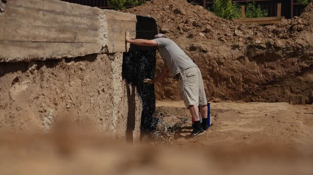 Professional construction worker applying a black bitumen waterproofing membrane to the concrete foundation wall of a building in an excavated trench during a sunny day at the construction site