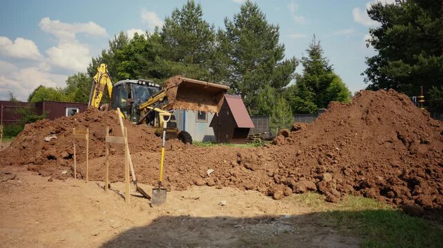 Heavy construction machinery working on a building site, with an excavator digging soil and moving earth to prepare the ground for foundation work during a sunny day with blue sky and trees