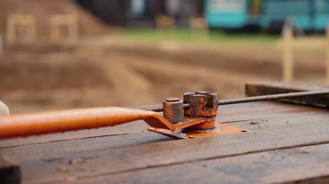 Muscular builder wearing protective gloves using a manual bending tool to shape a steel rebar on a wooden table at a construction site, preparing reinforcement for a concrete foundation