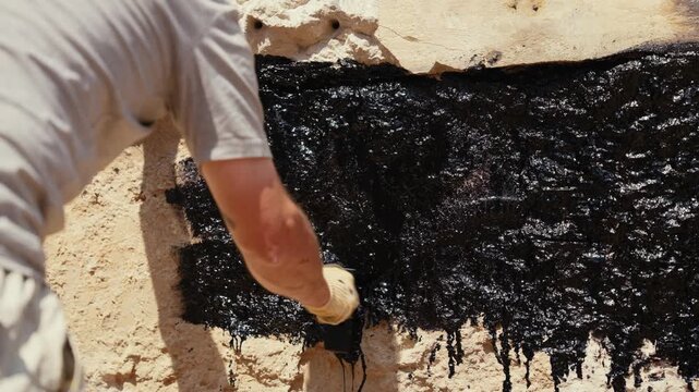 Professional construction worker applying a thick layer of black bitumen waterproofing tar to an exterior foundation wall with a brush, creating a protective sealant against moisture