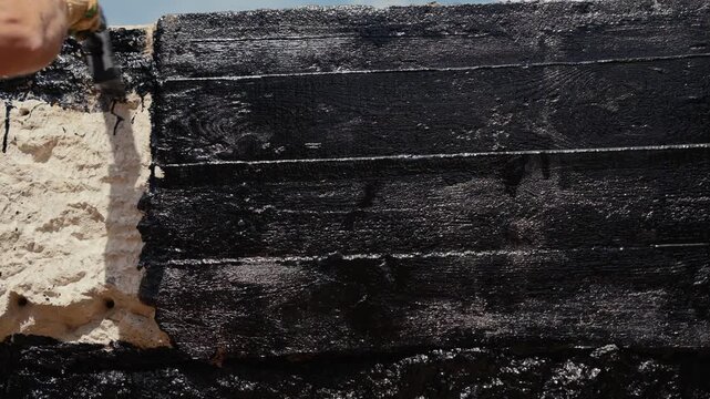 Professional builder applying a thick layer of black bitumen mastic to the foundation of a house, using a brush for waterproofing and sealing the concrete and wood surface against moisture