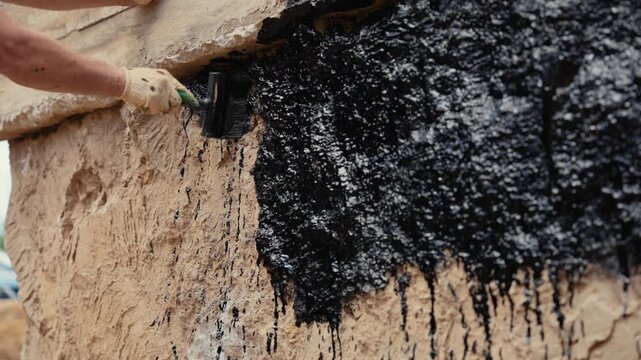 Professional builder wearing protective gloves applying black waterproofing tar to a building foundation with a wide brush, protecting the structure from water damage during construction