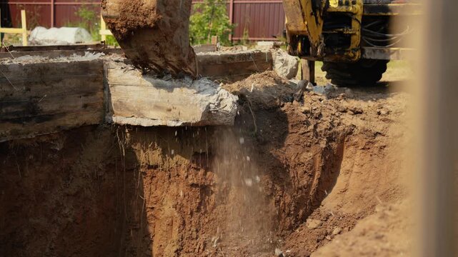 Heavy machinery with a bucket attachment demolishing an old concrete foundation at a construction site, with dirt and dust falling into the excavated pit during the dismantling process