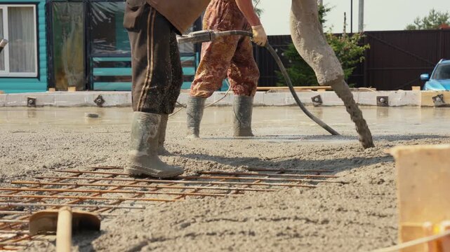 Unrecognizable builders pouring wet cement mixture with a concrete pump hose and leveling the slab with a vibrator on a reinforced steel rebar grid at a new construction site