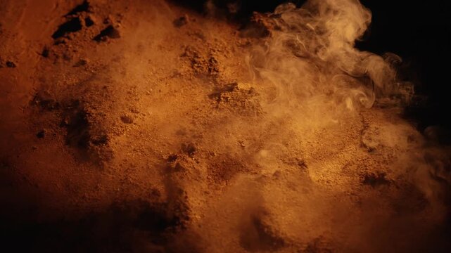 Mysterious white smoke billowing across a desolate, sandy surface with dramatic lighting. The warm, orange tones create an abstract and atmospheric scene against a dark, shadowy background
