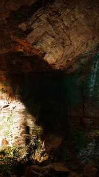 Inside a cave, sunlight filters through crevices, creating shadows on rocky walls. Plants grow at the cave floor, showing life amidst the stone. This scene captures natural beauty and light.
